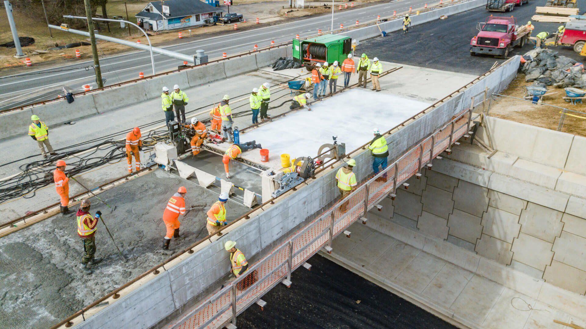Aerial view of a construction crew in high-visibility vests and hard hats pouring and finishing a concrete bridge deck over a road, with construction equipment, traffic cones, and vehicles visible along the adjacent highway.